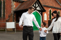 Smiling childholding the hands of their smiling parents outside of a church building