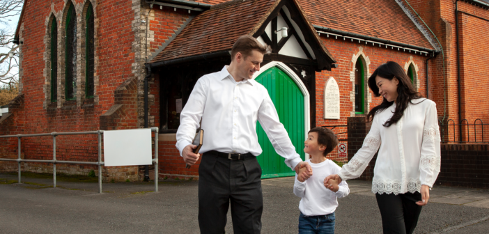 Smiling childholding the hands of their smiling parents outside of a church building
