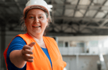 Construction worker in a hard hat and orange safety vest reaching out their hand to you in a room under construction