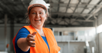 Construction worker in a hard hat and orange safety vest reaching out their hand to you in a room under construction